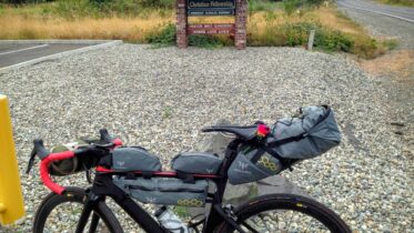 A touring bike fully equipped with gear bags, parked beside a gravel roadside near a rustic sign for Summit Christian Fellowship. The sign and surrounding overgrown grass set against a road backdrop evoke a sense of a remote or rural location, hinting at long-distance cycling adventures.