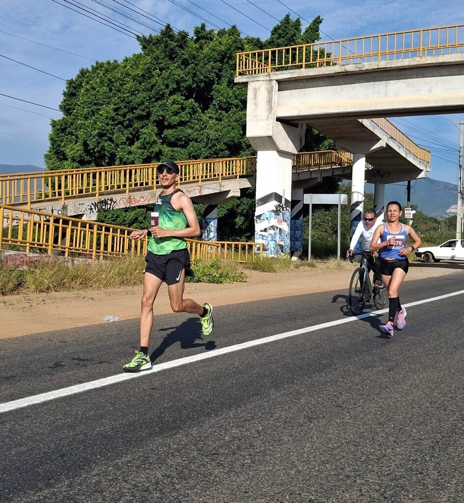 Oax Sport runner wearing bib 566 runs ahead of a woman in blue while Arnulfo follows on a bicycle during the Oaxaca marathon.