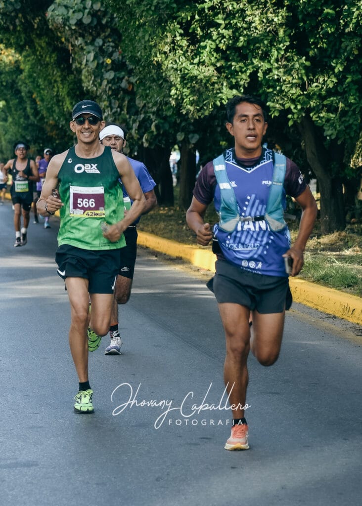 Oax Sport runner wearing bib 566 runs alongside another athlete on a tree-lined section of the Oaxaca marathon.
